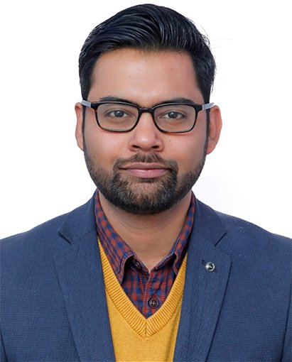 Portrait of a man with dark hair, beard, and glasses, wearing a navy blazer over a yellow sweater and plaid shirt against a white background.